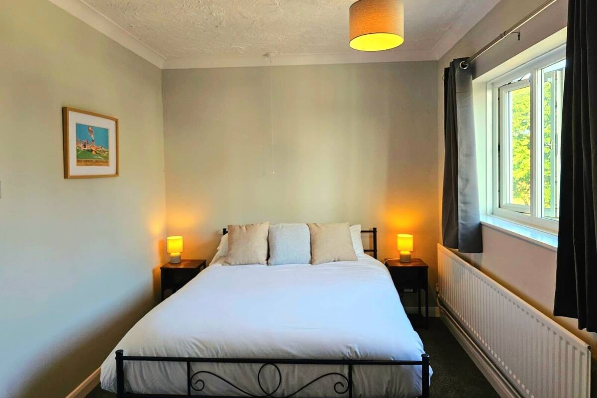 Une chambre à coucher comprenant un lit, deux lampes de chevet, un luminaire au plafond, une fenêtre avec des rideaux et un radiateur.