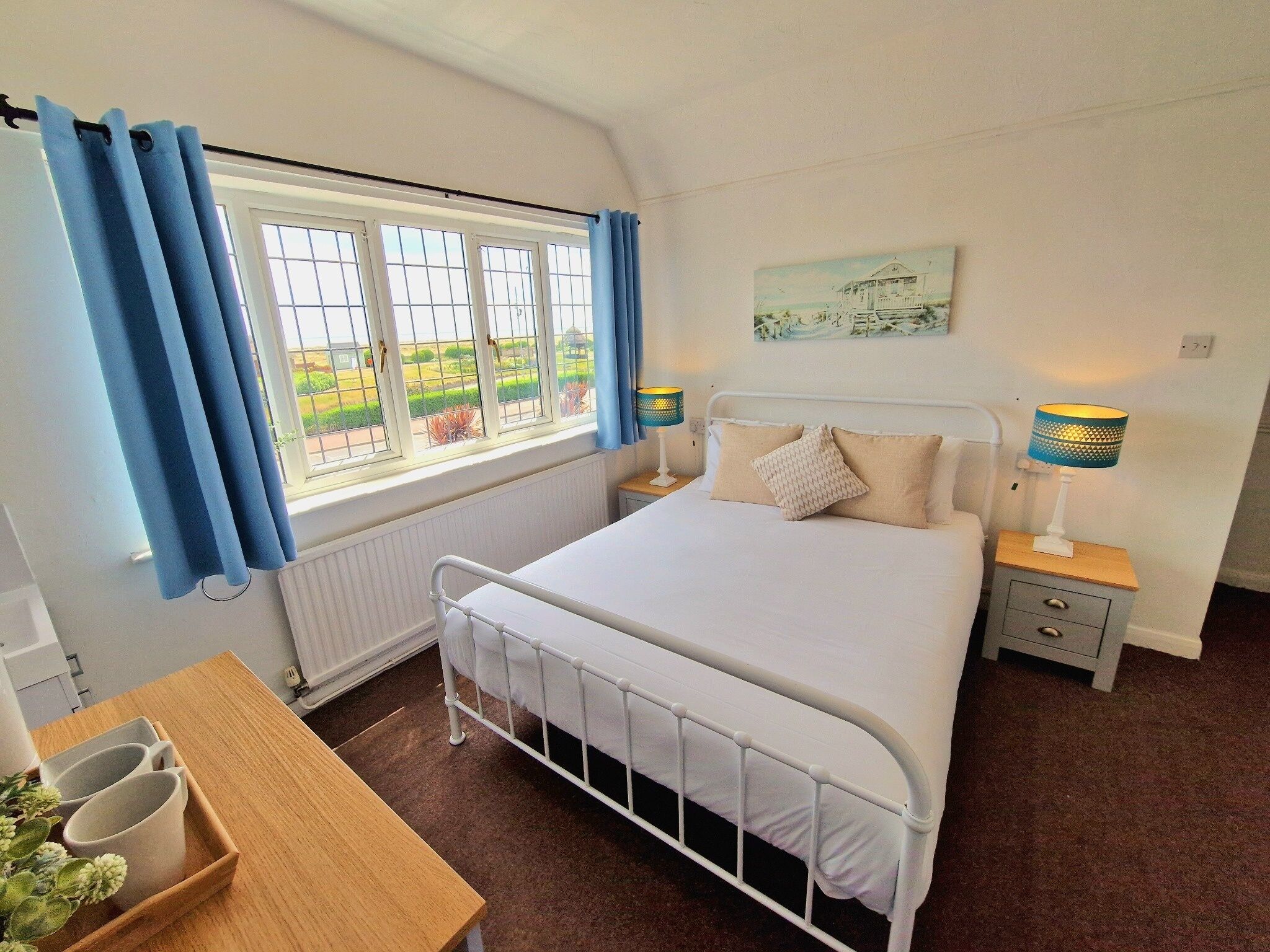 A bedroom with a bed, bedside tables, a window with curtains, a radiator, and a small table with cups.