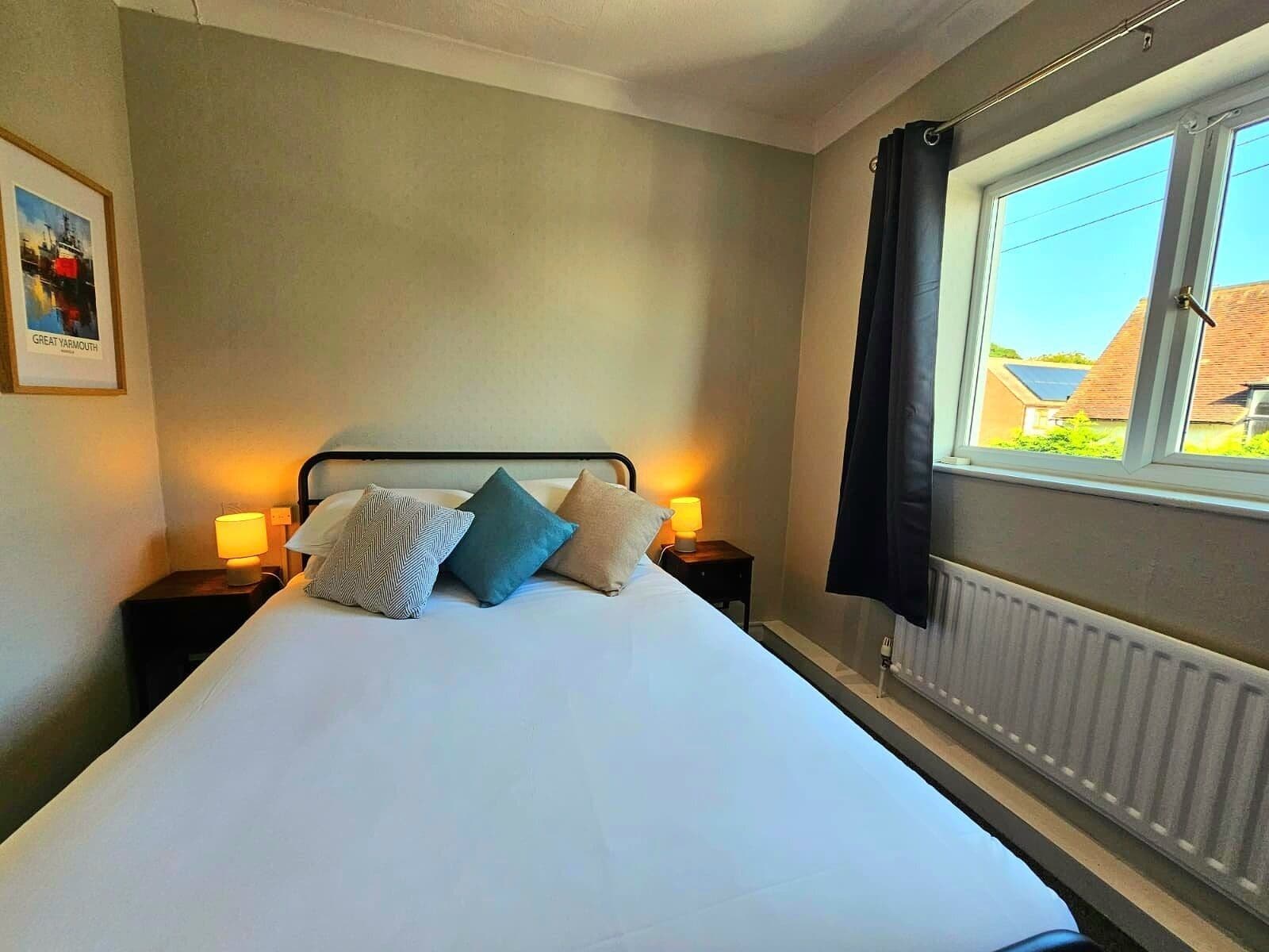 A neatly made bed with three pillows, two bedside tables with lamps, a framed picture on the wall, and a window with curtains.