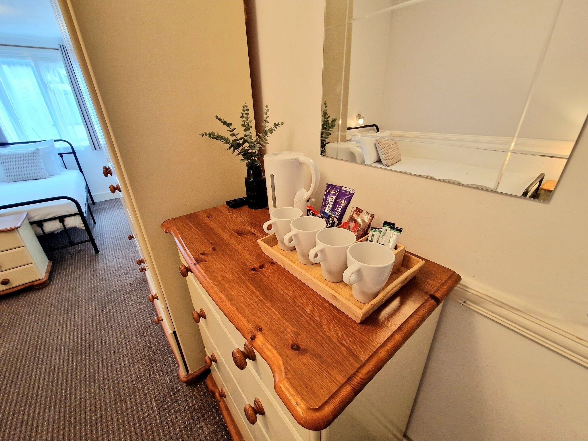 A wooden dresser with a tray holding white mugs and coffee packets, a small plant, and a mirror above it.