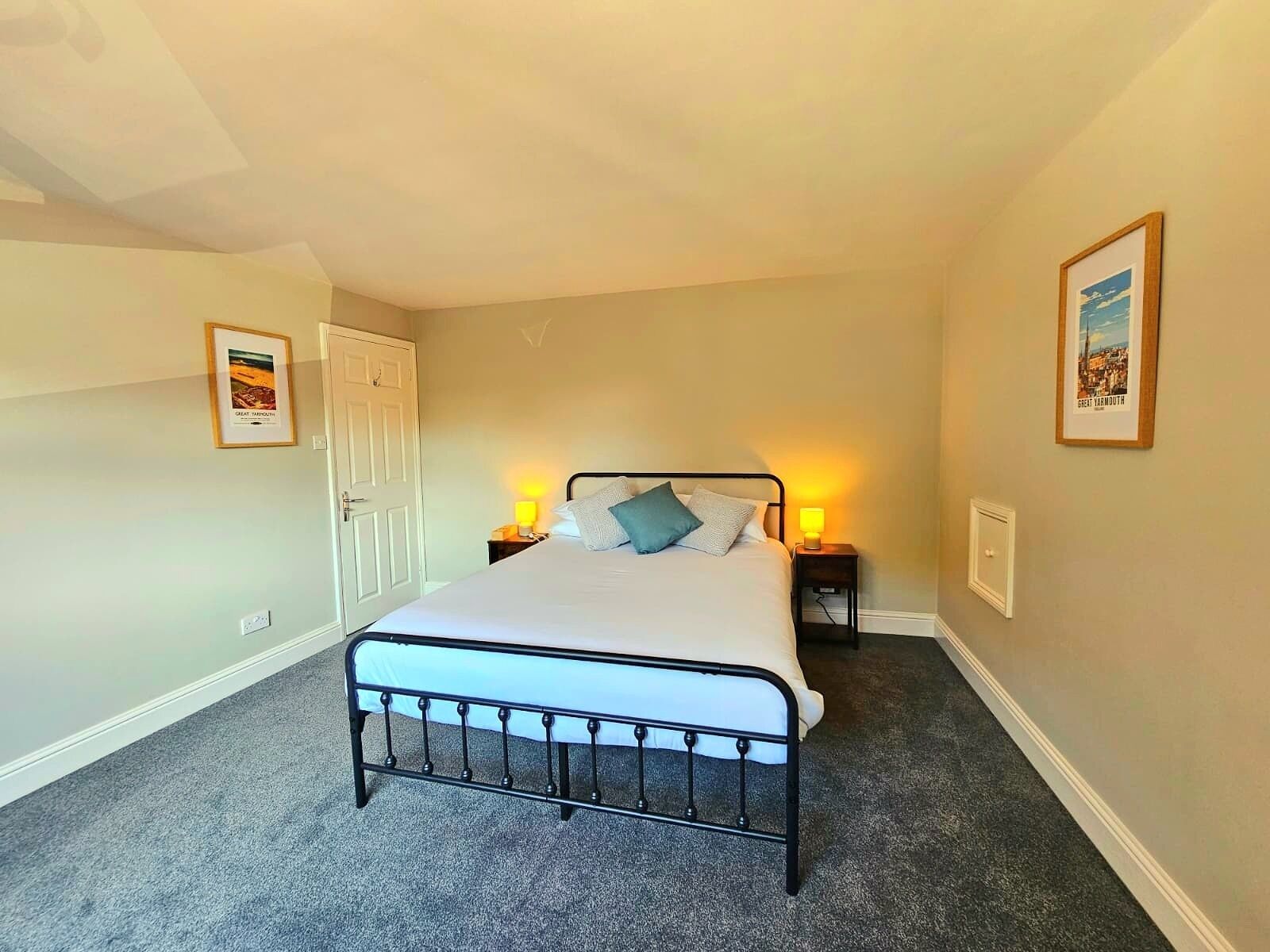 A bedroom with a single bed, two bedside lamps, a door, and two framed pictures on the walls.
