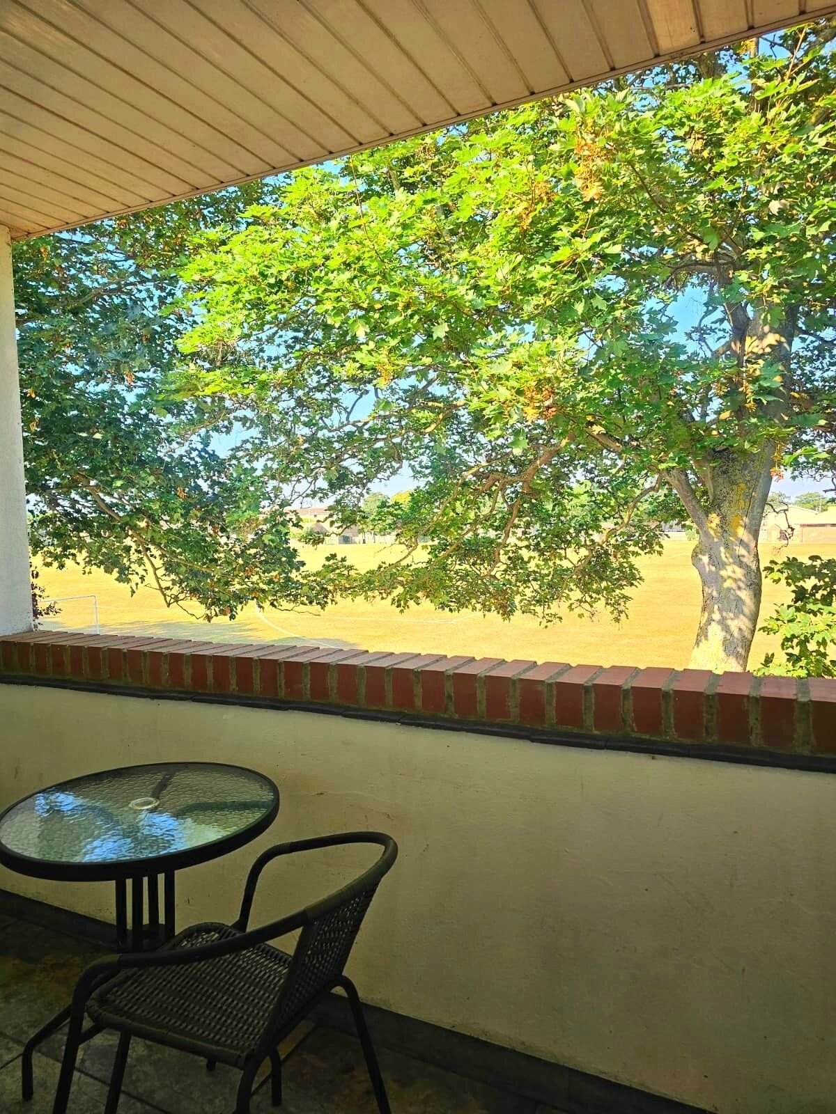 A covered outdoor area with a table and chairs, a brick wall, and a view of a tree and open space.