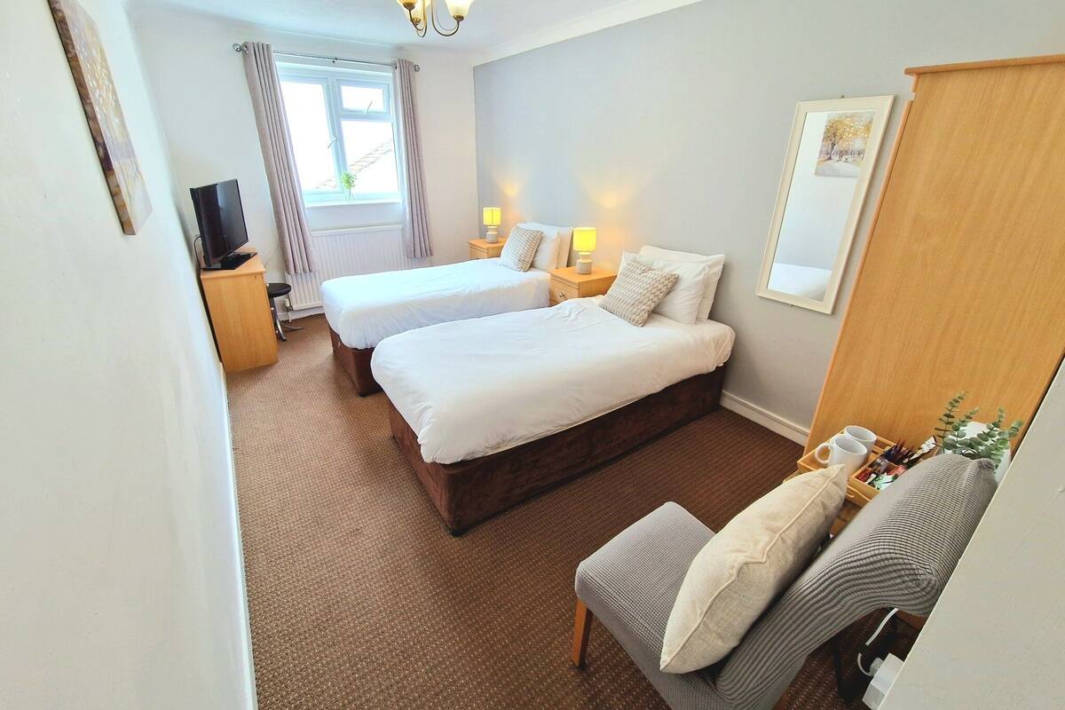 Une chambre d’hôtel avec deux lits, une télévision, une fenêtre avec des rideaux, une armoire en bois et une petite table avec une lampe.