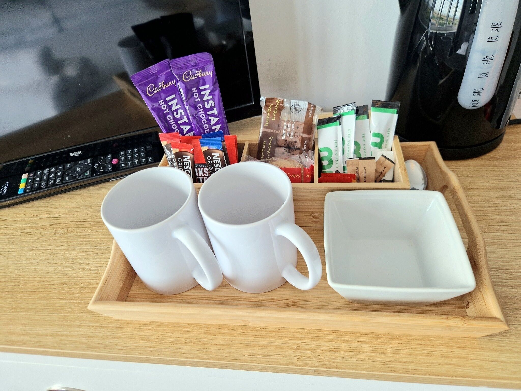 A tray with two white mugs, a white bowl, and assorted snack packets including Cadbury and Nescafé.