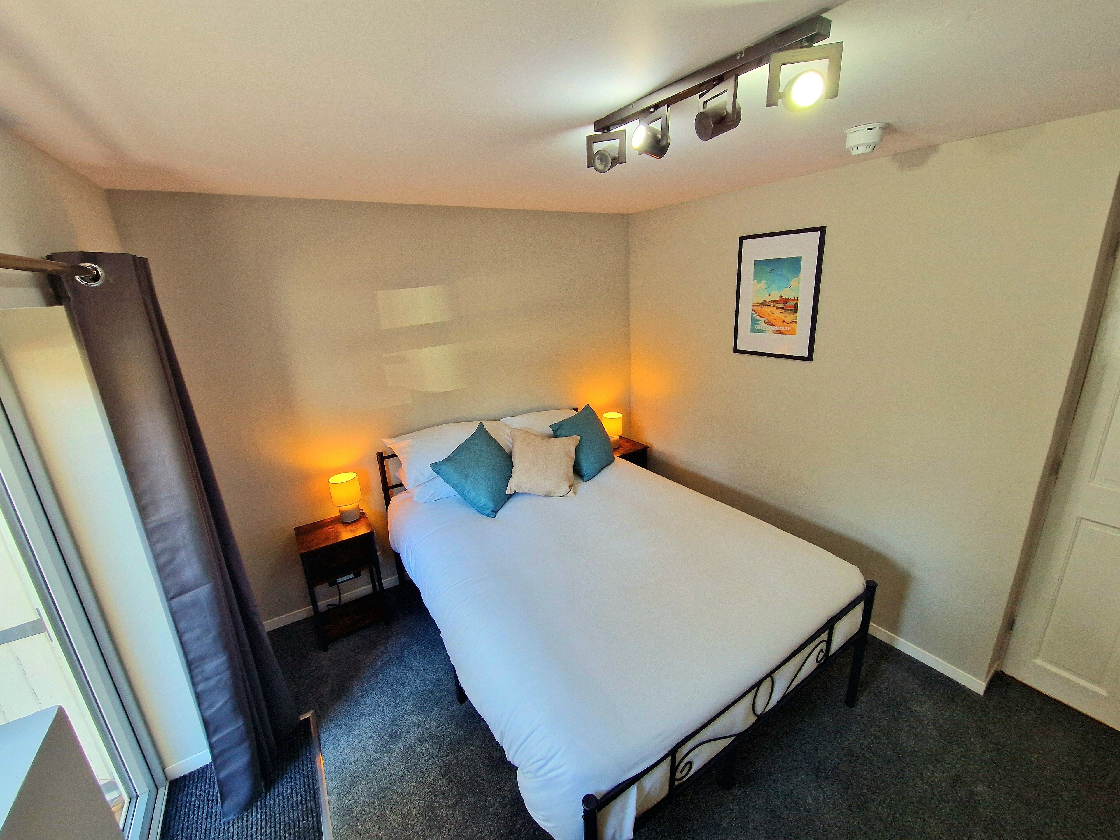 A neatly made bed with white linens and blue pillows, a bedside lamp, and a framed picture on the wall.