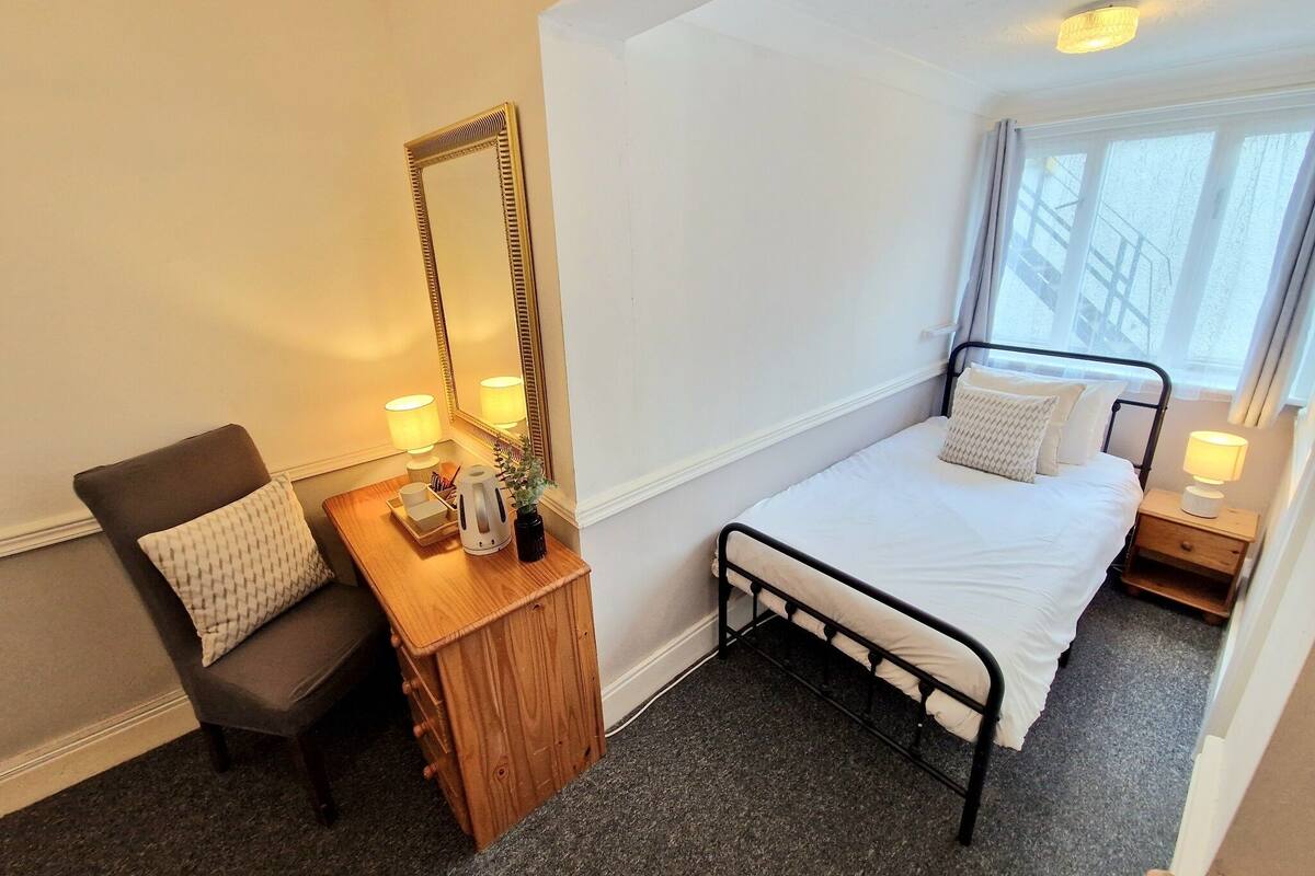 Une chambre à coucher avec un lit, une chaise, une table de chevet en bois, un miroir et une fenêtre avec des rideaux.