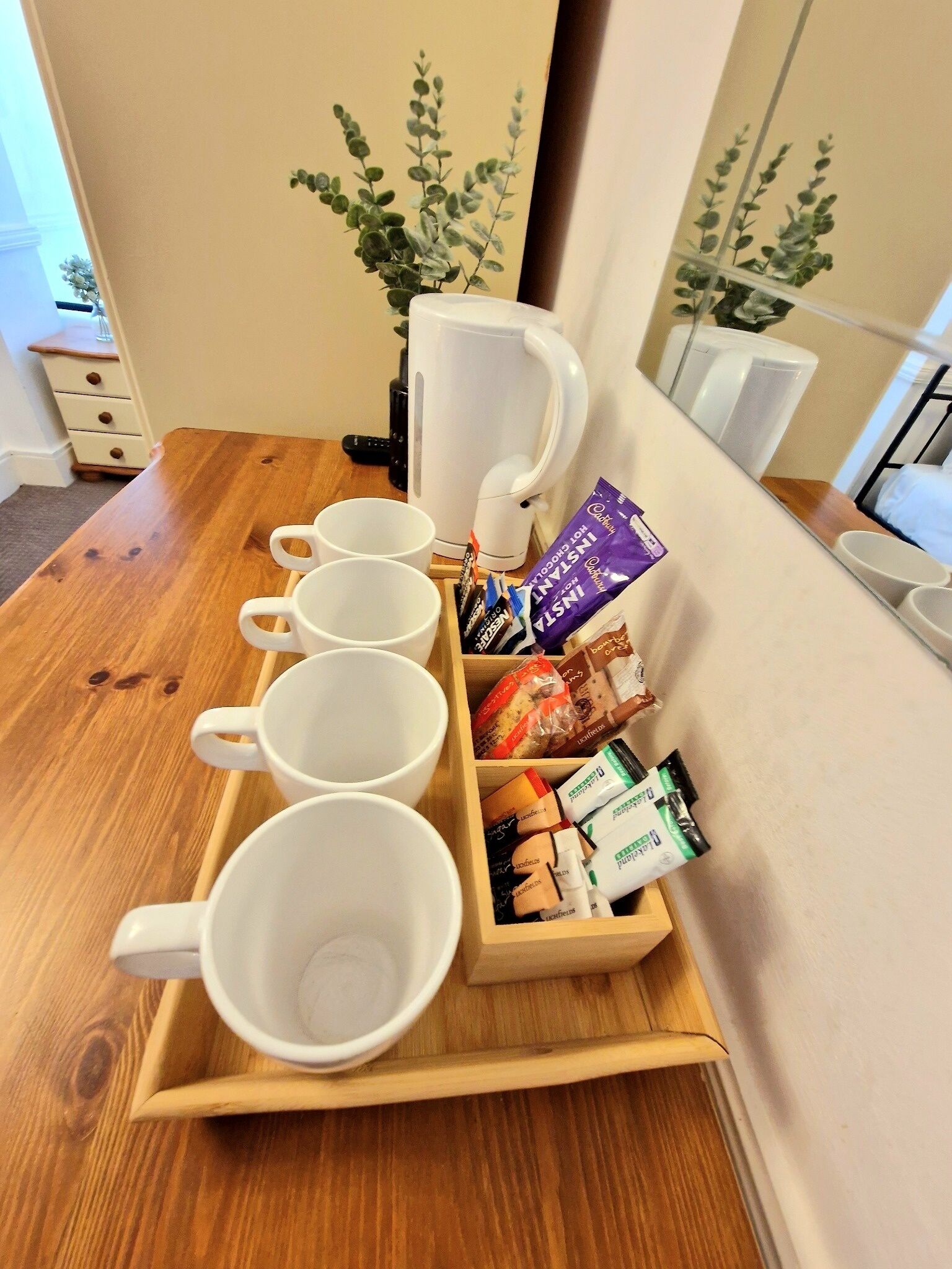 A wooden tray with white mugs and snacks, a white kettle, and a potted plant in a hotel room.