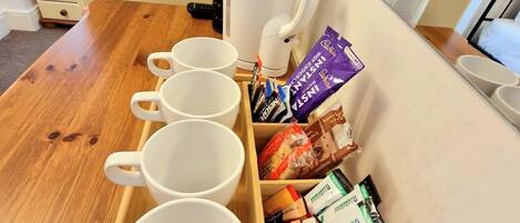 A wooden tray with white mugs and snacks, a white kettle, and a potted plant in a hotel room.