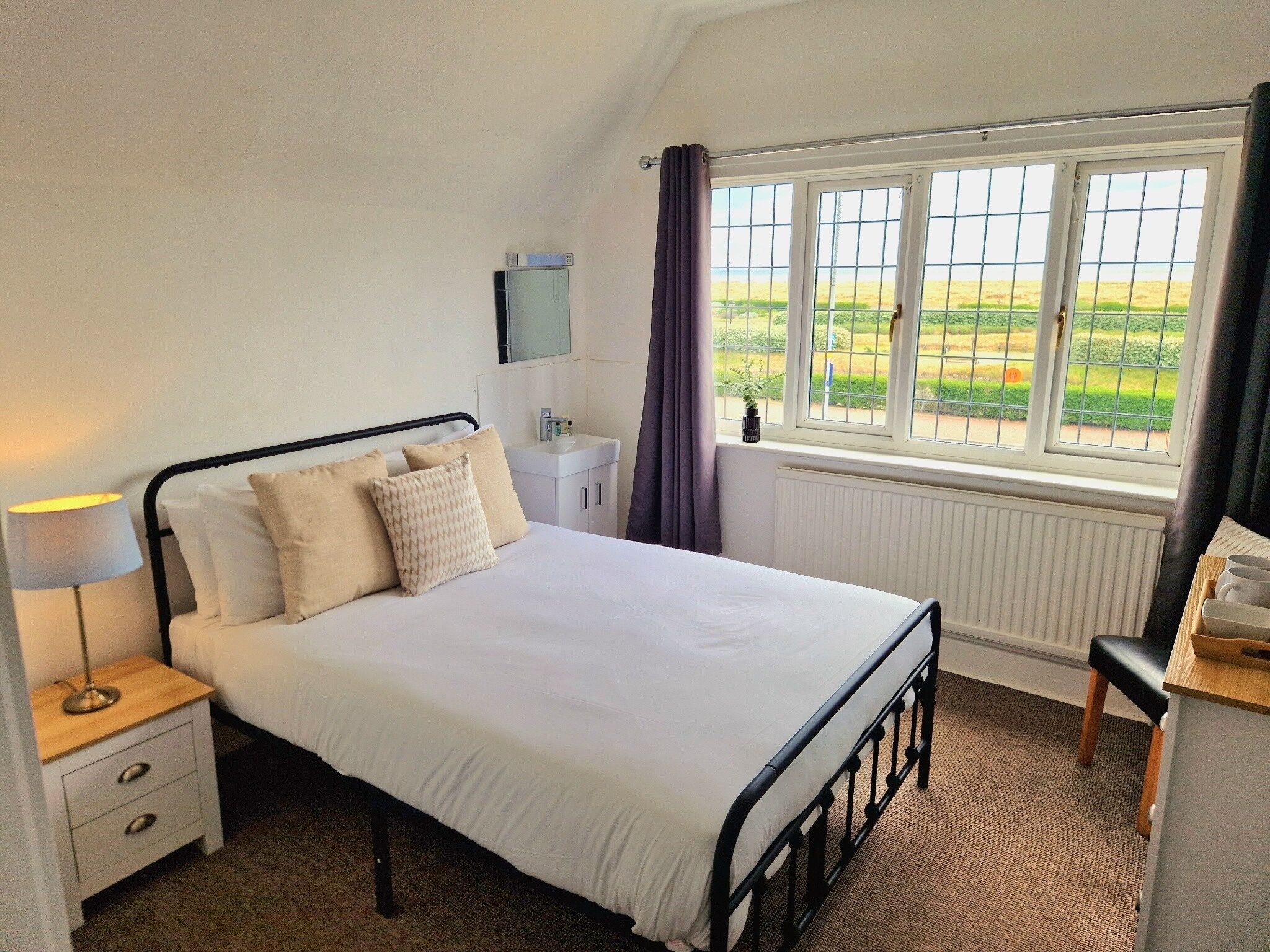 A bedroom with a bed, bedside table, lamp, and a window with a view of the outdoors.