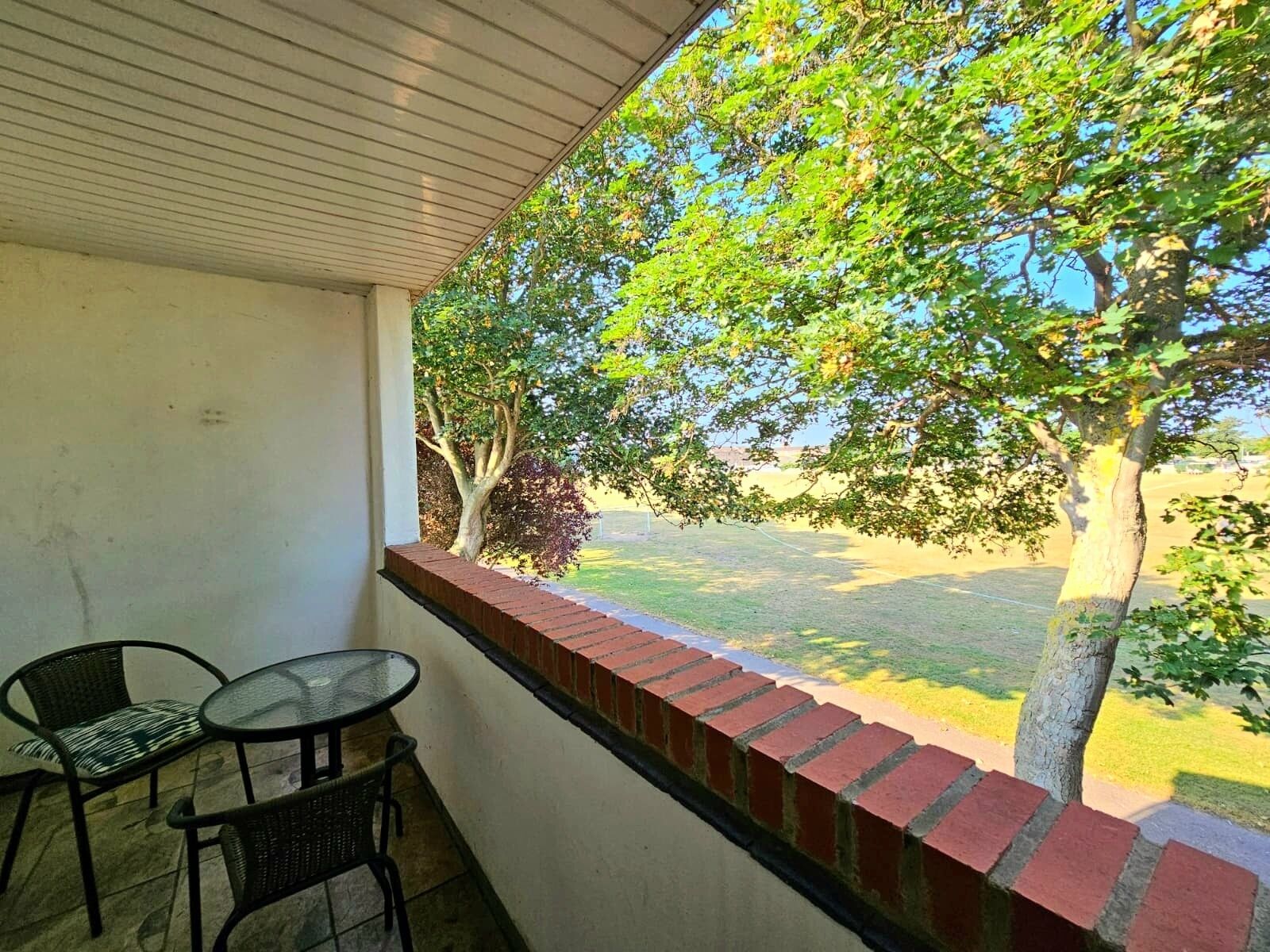 Un balcon abrité, doté d’une table et de chaises, un mur en briques et une vue sur un champ verdoyant et des arbres.