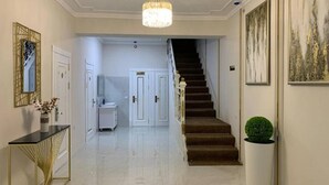 A hallway with a staircase leading to a second floor. There is a console table with a vase of flowers, a chandelier, and framed artwork on the walls. - Central Plaza (Urgench)