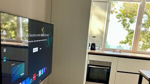 Una cocina moderna con un televisor de pantalla plana mostrando diversas aplicaciones de transmisión, un banco de madera con un tazón y recipientes con forma de corazón, y un ventanal con vistas a árboles.