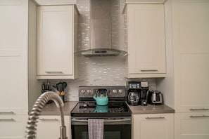 Una cocina moderna con armarios blancos, una campana extractora de acero inoxidable, una tetera azul, una cafetera y un horno empotrado.