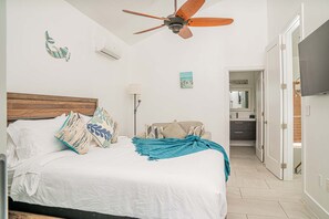 Un dormitorio con cama, ventilador de techo, aire acondicionado de pared, lámpara de noche y vista al baño.