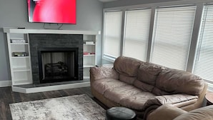 A modern living room with a beige leather sofa, a flat-screen TV mounted above a fireplace, and a large window with blinds.
