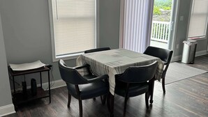 A dining area with a table and four chairs, large windows with blinds, and a view of greenery outside.