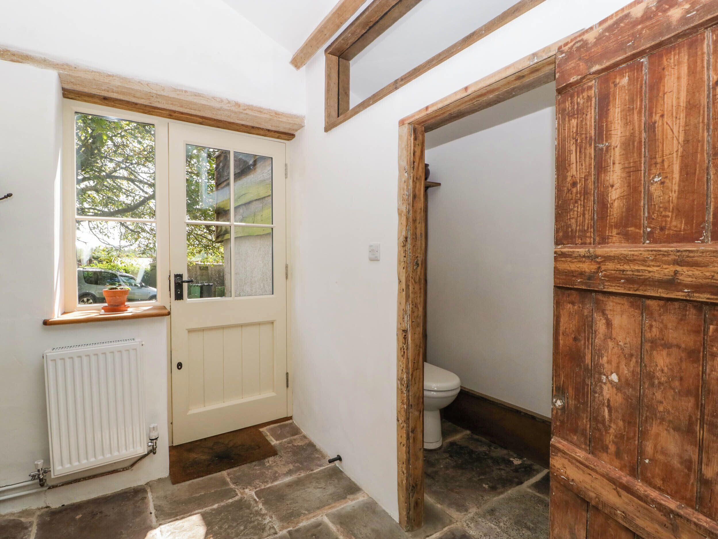 A narrow hallway with a white door, a window with a view of the outside, a radiator, and a wooden door leading to a bathroom.