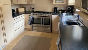 A modern kitchen with light wood cabinets, a stainless steel stove, a built-in oven, a black countertop, a sink, and a dish rack.