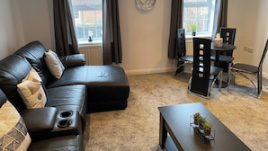 A modern living room with a black leather sofa, a dining table with chairs, and a large window with curtains.