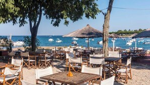 An outdoor seating area with tables and chairs, a thatched umbrella, and a view of the sea with boats. - Camping Park Umag - Eurocamp (Brtonigla)