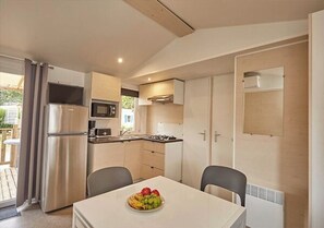 A compact kitchen with a refrigerator, microwave, and stove. There is a dining table with chairs and a fruit bowl. - Camping Park Umag - Eurocamp (Brtonigla)