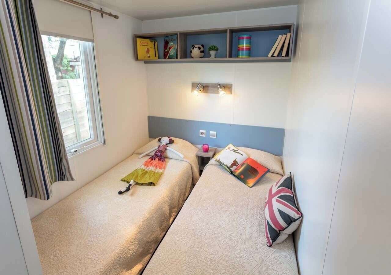 A compact room with two beds, a window with curtains, a shelf with books and decorative items, and a bedside table with a lamp.