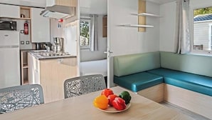 A compact kitchenette with a microwave, stove, and sink. There is a dining table with a plate of bell peppers. A green cushioned bench is against the wall, and a window with curtains is visible. - Camping Park Umag - Eurocamp (Brtonigla)
