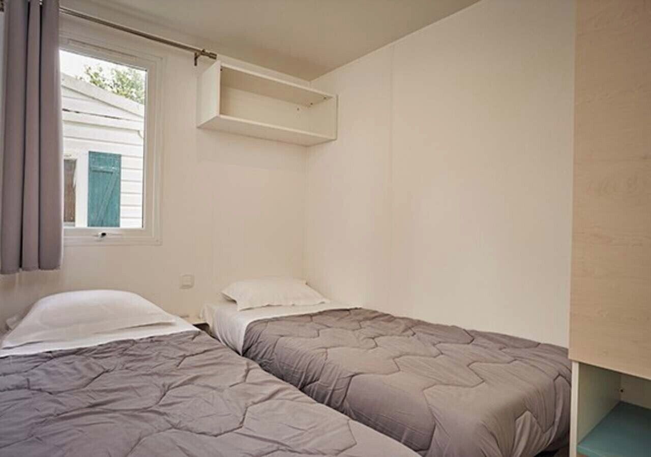 Een kleine, eenvoudige kamer met twee eenpersoonsbedden, een raam met gordijnen en een ingebouwd plankje boven de bedden.