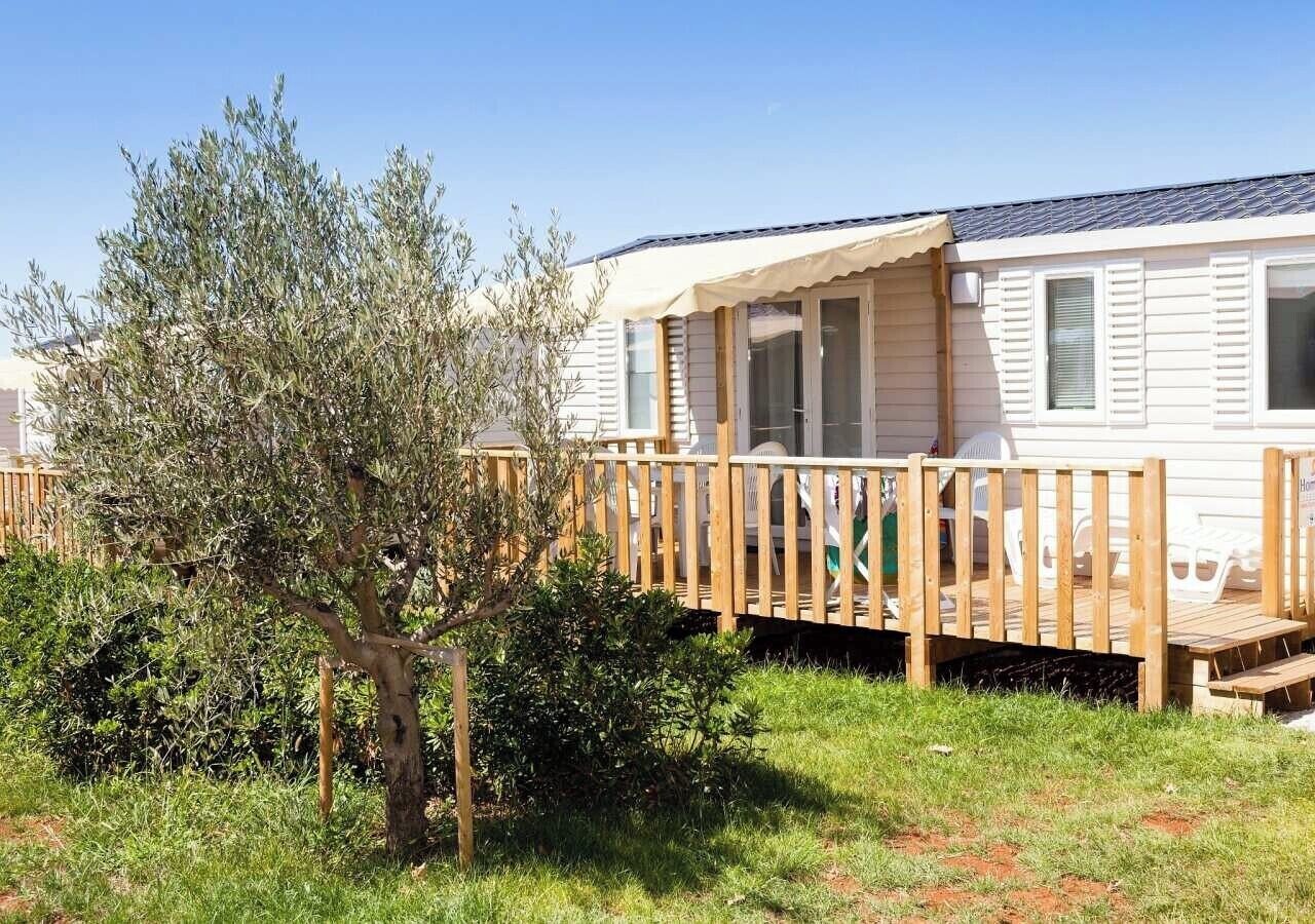 A mobile home with a wooden deck, a covered porch, and a small garden with a tree and shrubs.