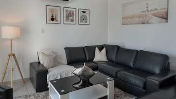 Un salon moderne avec un canapé en cuir noir, une table basse en verre et une lampe. Trois œuvres d’art encadrées ornent le mur.