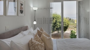 A bedroom with a bed, pillows, a nightstand, a lamp, and a view of greenery outside. - Phaedrus Living: Seaview Hilltop Maisonette (Chlorakas)