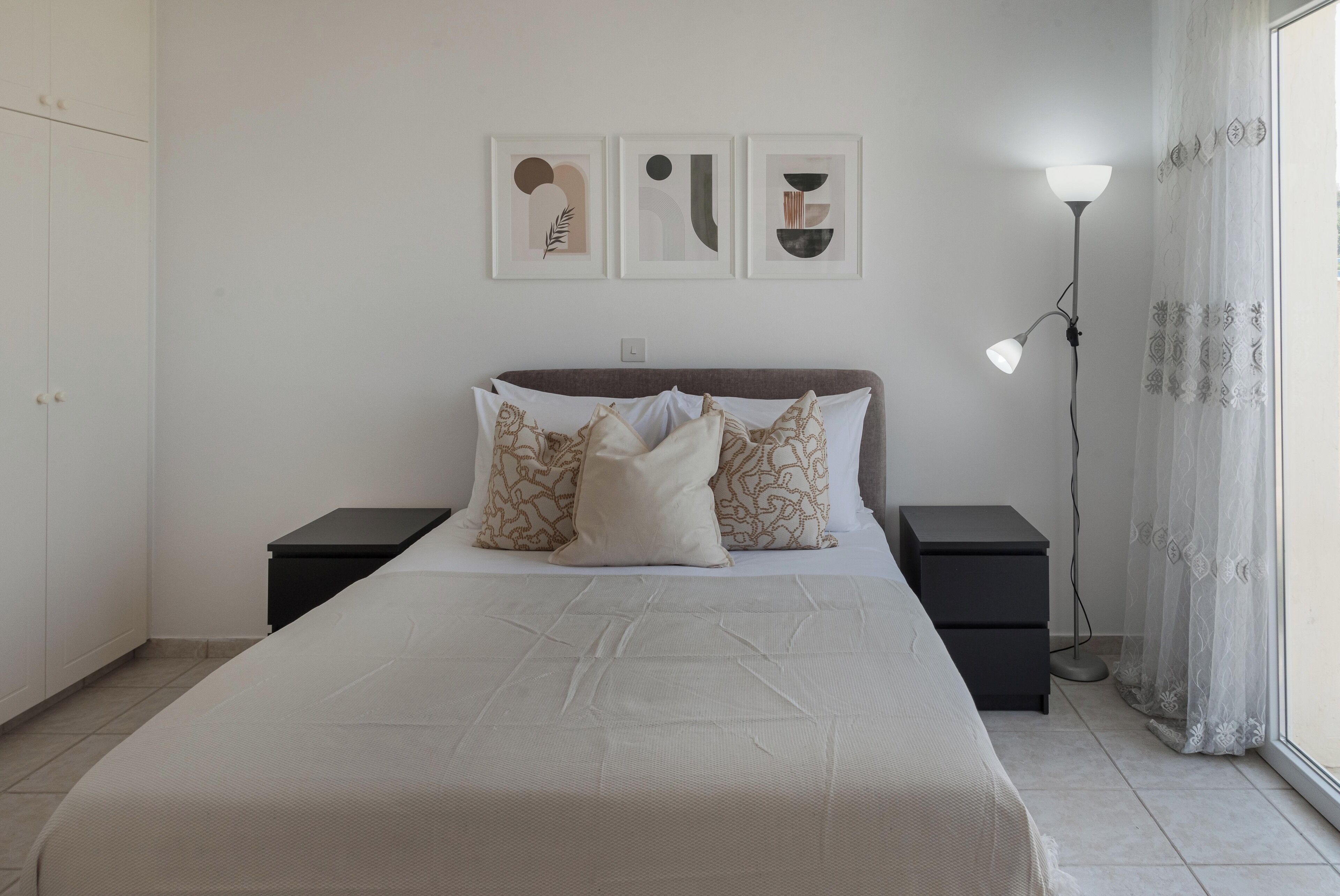 A bedroom with a large bed, two bedside tables, a floor lamp, and three framed artworks on the wall.