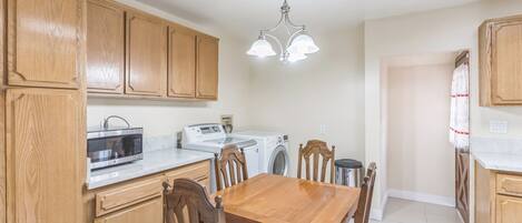 A kitchen with wooden cabinets, a microwave, a washing machine, and a dining table with chairs.