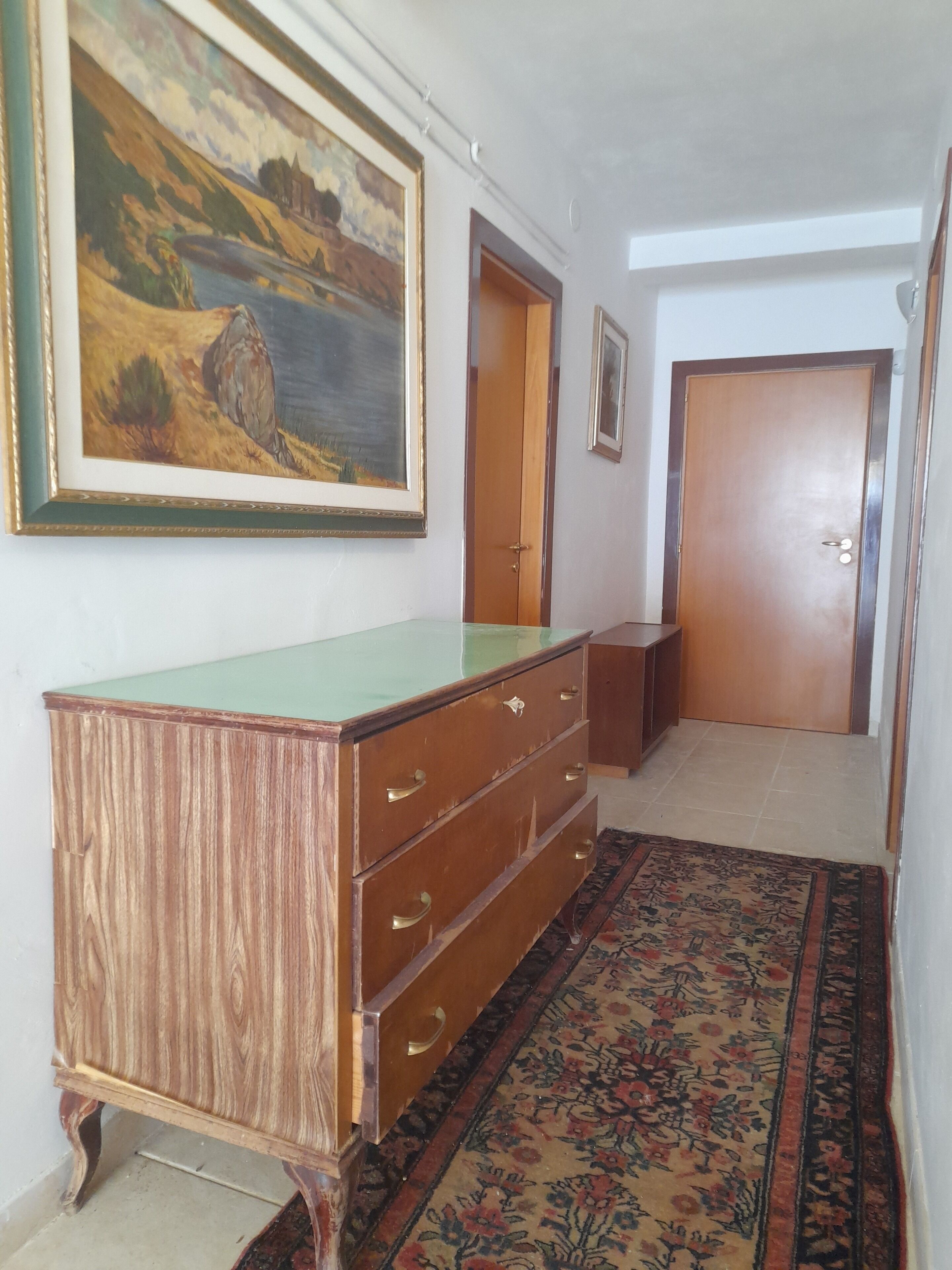 Un couloir avec une commode en bois, un tableau, une porte et un tapis à motifs.