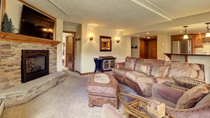 Ein Wohnzimmer mit Kamin aus Stein, einem großen Flachbildfernseher darüber, einem Ecksofa und einer Küchenzeile mit Holzausstattung und Pendelleuchten.