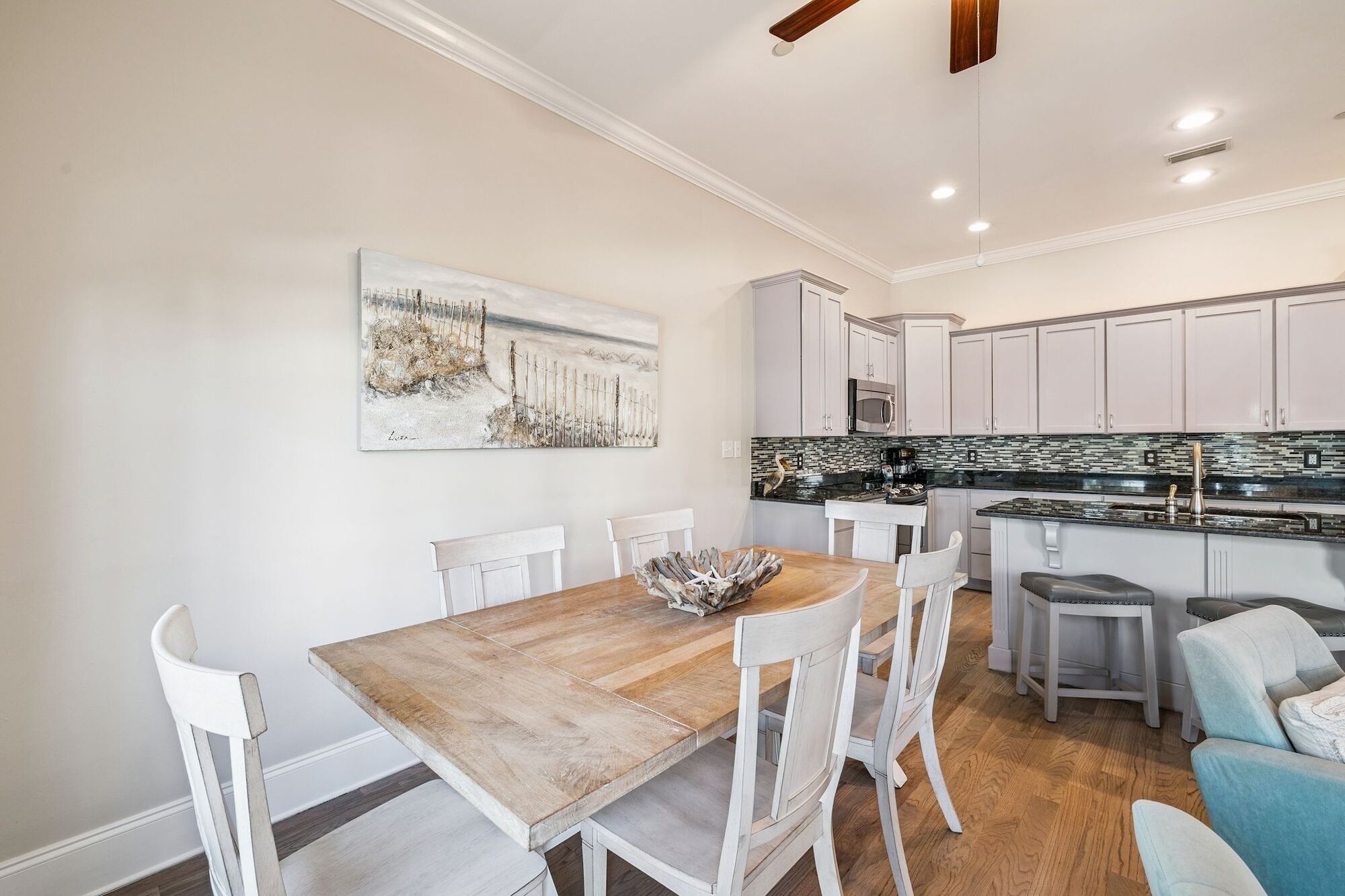 A dining area with a wooden table and white chairs, a kitchen with white cabinets and a black countertop, and a ceiling fan.