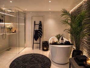 A modern bathroom with a glass shower enclosure, a freestanding bathtub, a black rug, and a potted plant.