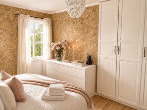 A bedroom with a bed, a dresser, a window with curtains, a chandelier, and a wardrobe.