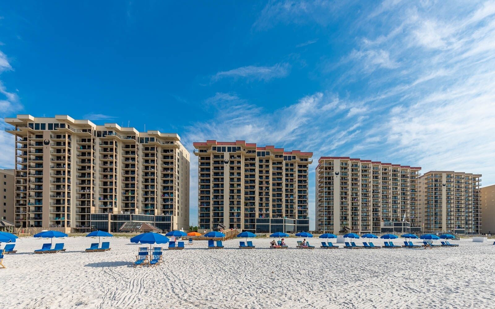A beachfront hotel with multiple high-rise buildings, a sandy beach with blue lounge chairs and umbrellas, and clear blue skies.