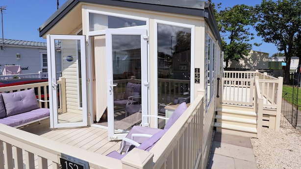 A small, single-story building with a deck, a covered seating area with cushions, and a number sign indicating its location. - 152 Central Park (Prestatyn)
