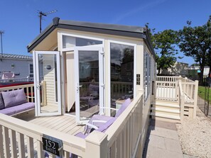 A small, single-story building with a deck, a covered seating area with cushions, and a number sign indicating its location. - 152 Central Park (Prestatyn)