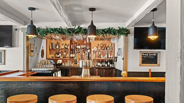 A bar area with wooden stools, a bar counter, shelves of bottles, a television, and a sign for drink specials.