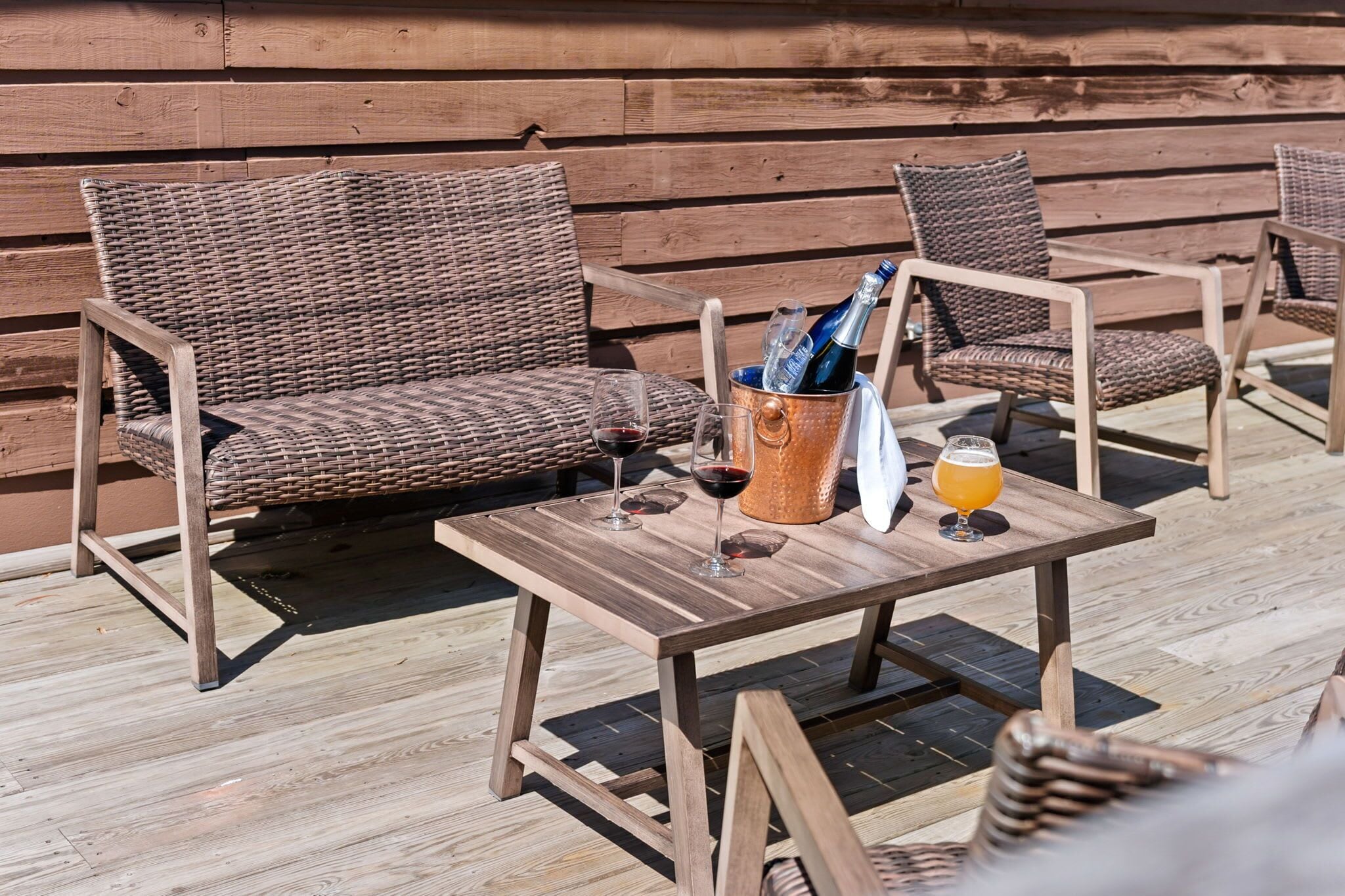 Des meubles en osier, une table en bois et un récipient contenant des bouteilles et des verres, le tout posé sur une terrasse en bois.
