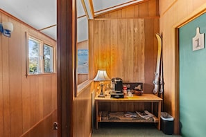 A compact room with wooden walls, a small table with a lamp, a coffee maker, and a ski rack.