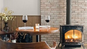 Un rincón comedor acogedor con pared de ladrillo, estufa de leña y dos copas de vino tinto sobre una mesa redonda.