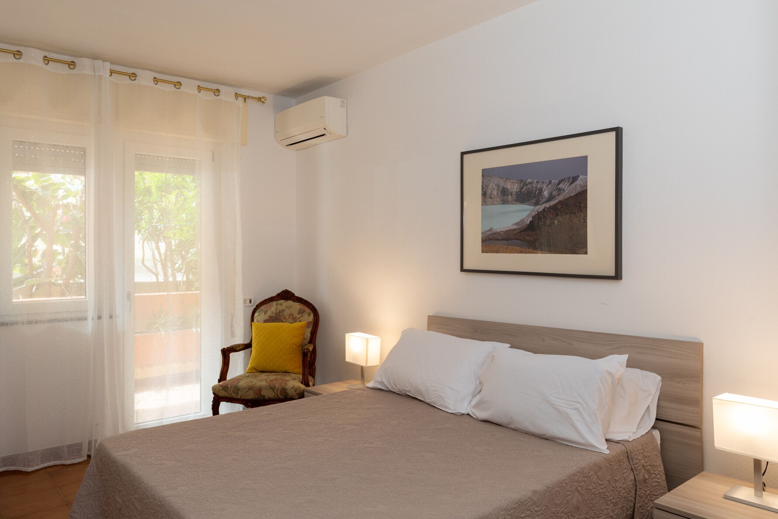 A hotel room with a bed, a chair, a nightstand, a lamp, an air conditioner, and a framed picture on the wall.