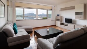 Una sala de estar moderna con un sofá, una mesa de centro y un televisor sobre un mueble bajo. Se ve un ventanal con vistas a un cuerpo de agua y montañas.