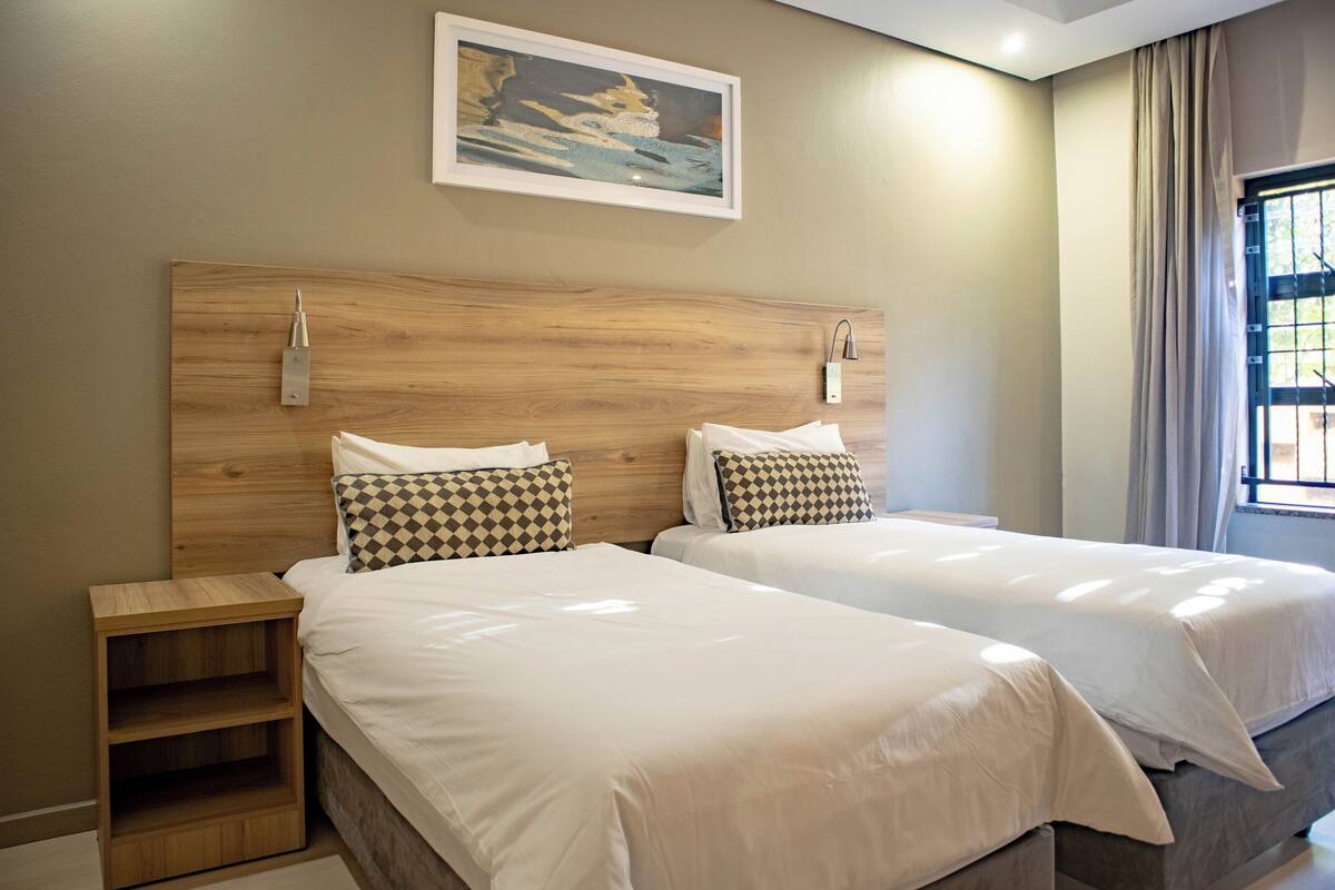Een hotelkamer met twee bedden, een houten hoofdbord, een nachtkastje, een schilderij aan de wand en een raam met gordijnen.