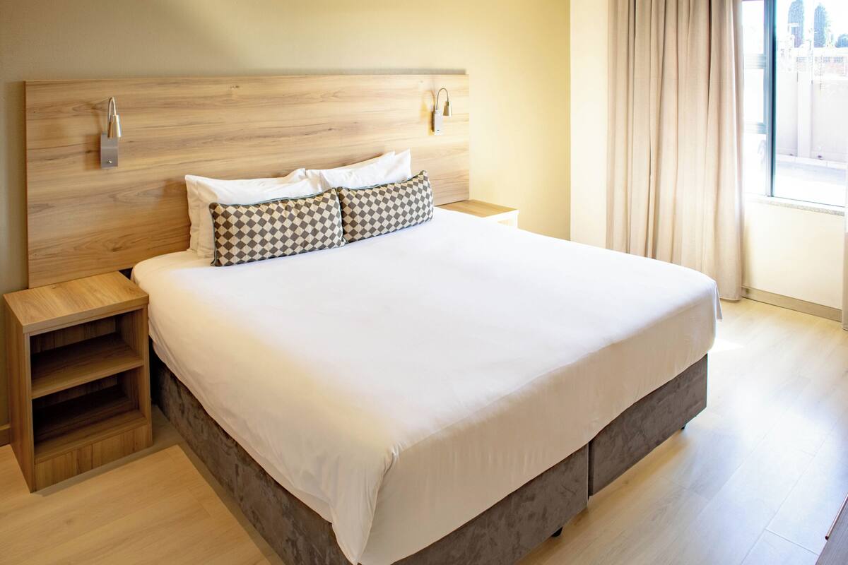 Een moderne hotelkamer met een houten hoofdbord, een groot bed met witte lakens, een nachtkastje van hout en uitzicht op de stad door het raam.