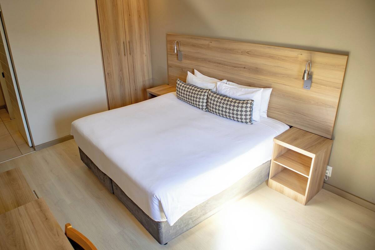 Een moderne slaapkamer met een houten hoofdbord, een net opgemaakt bed met witte lakens en een klein houten nachtkastje met lade.